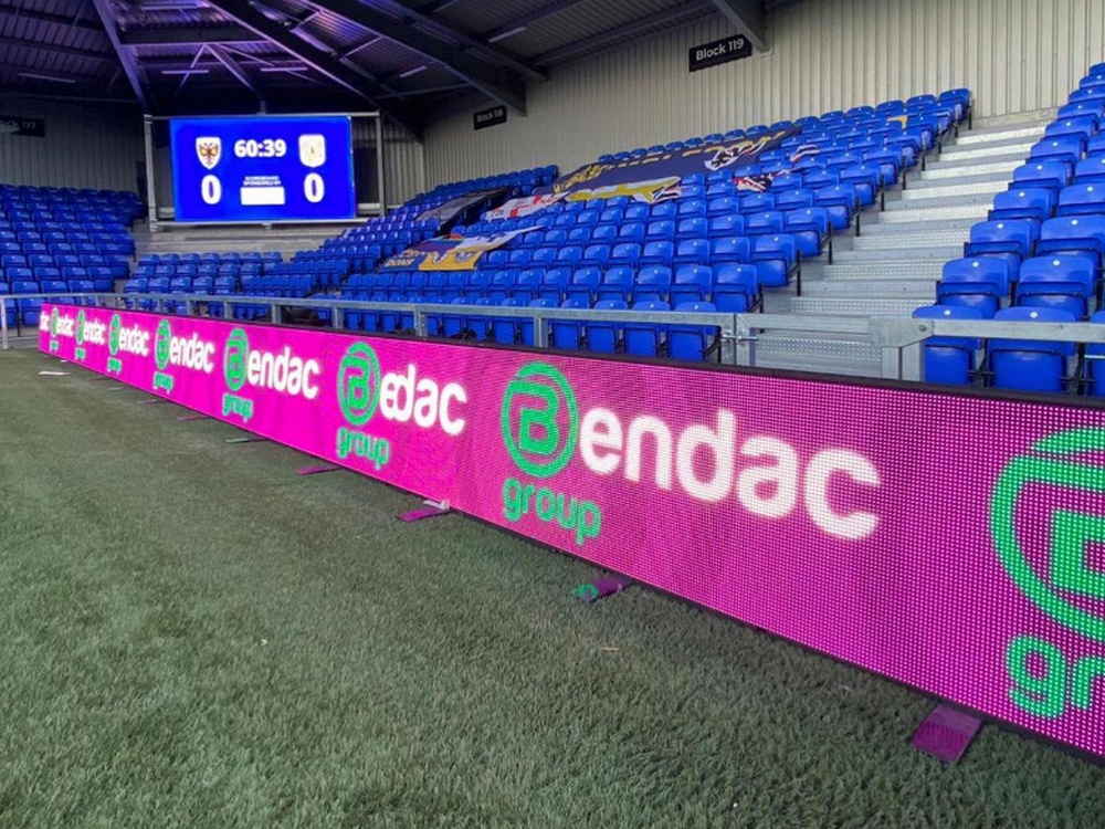 Blue football stadium seats with pink Bendac branding boards running around the edge of the pitch