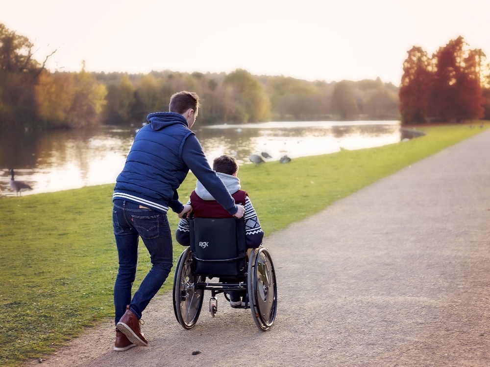 A young man pushes a boy in a wheel chair along a path next to a river