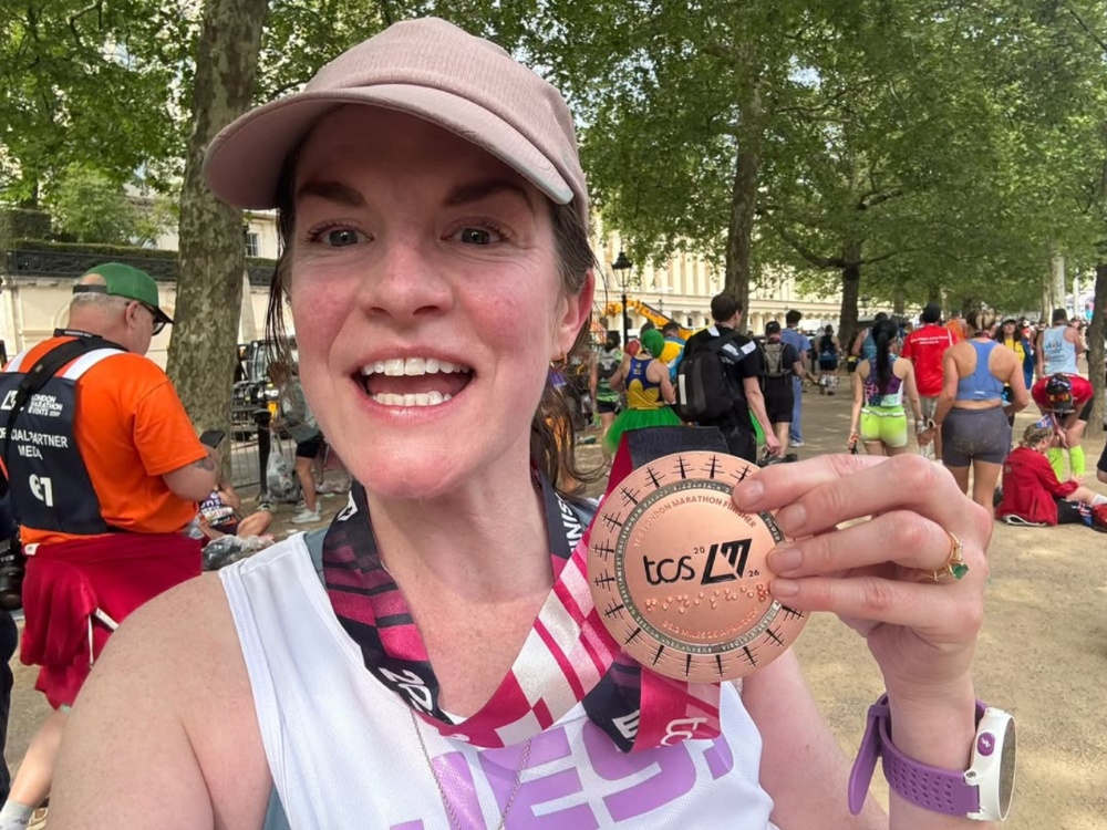A lady in a baseball cap smiling and holding her London marathon medal up