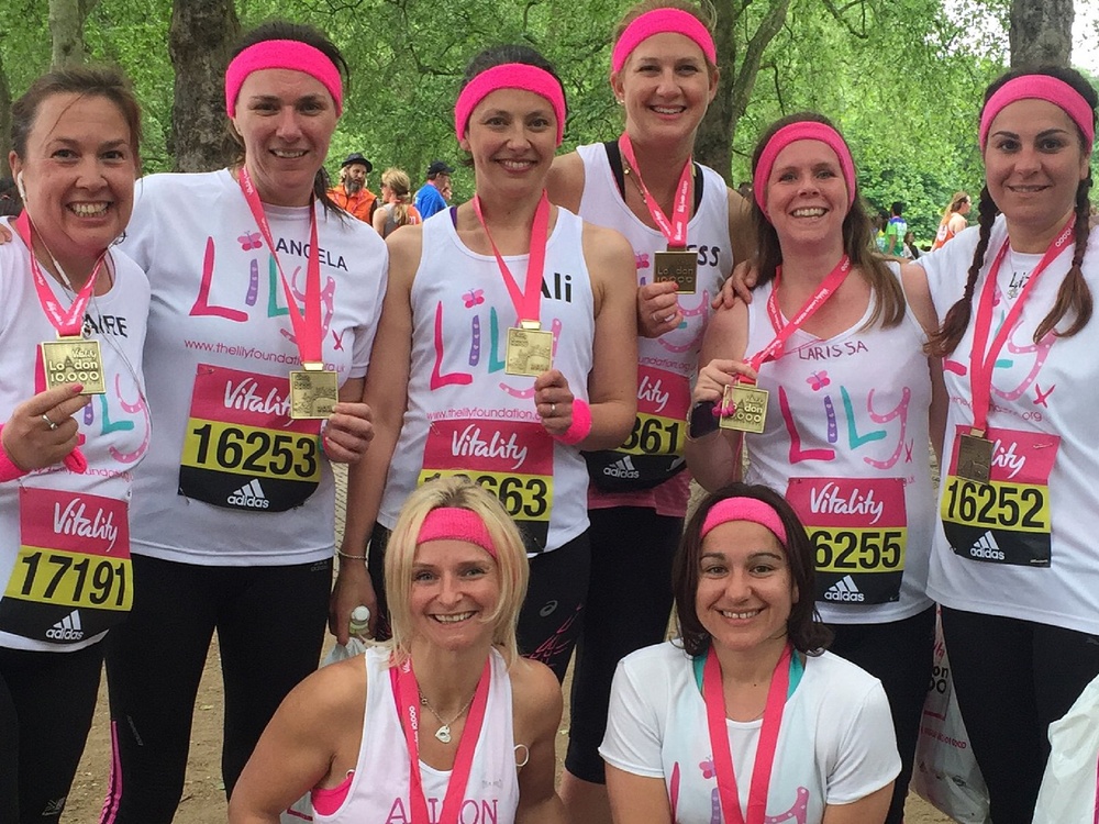A group of women in white lily running tops stand together wearing pink head bands wearing medals