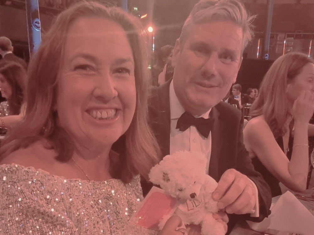 Lily team member Claire at a table with Sir Keir Starmer holding a Lily Share a Bear and smiling