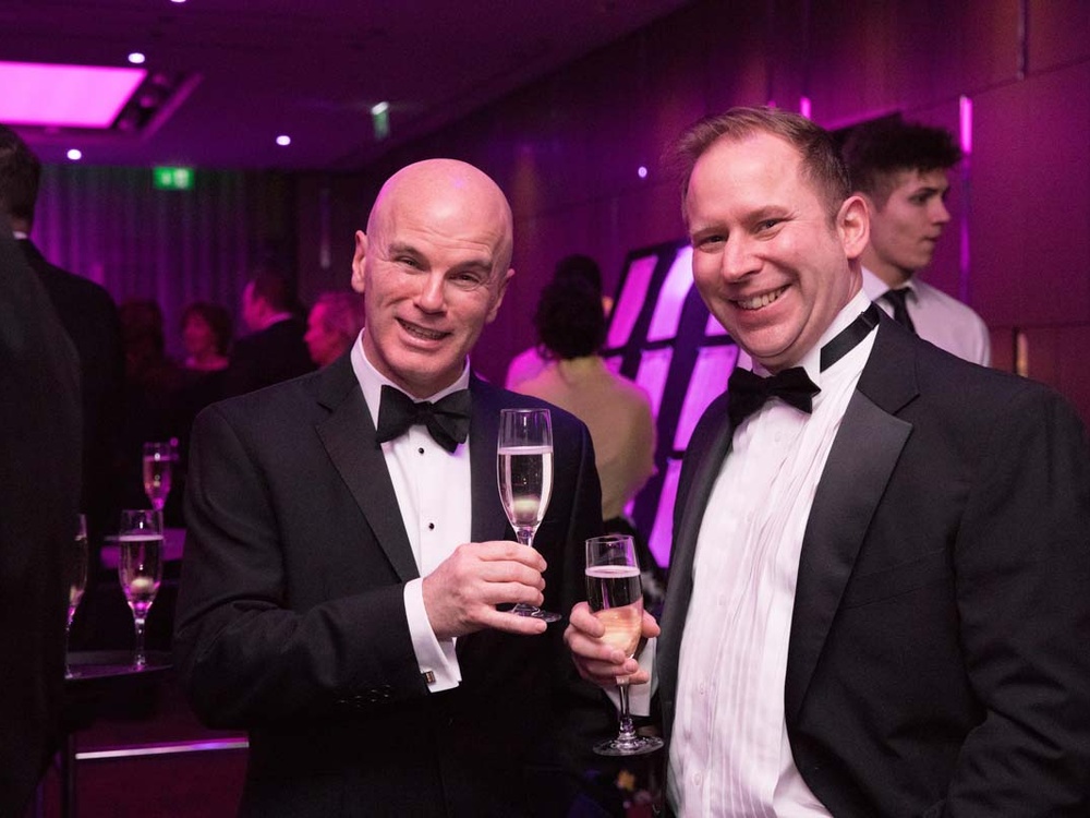 Two men in black tuxedos smiling and holding glasses of sparkly