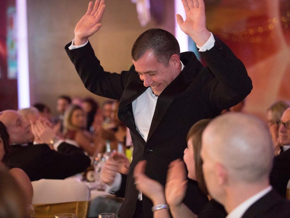 A man in a black tuxedo standing up laughing and raising his hands