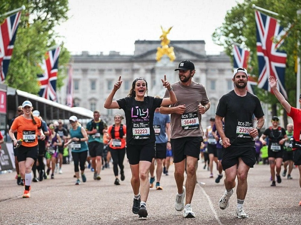 Real stories. Real support. Made possible by you. - A lady in a Lily top waving as she reaches the end of the London Marathon