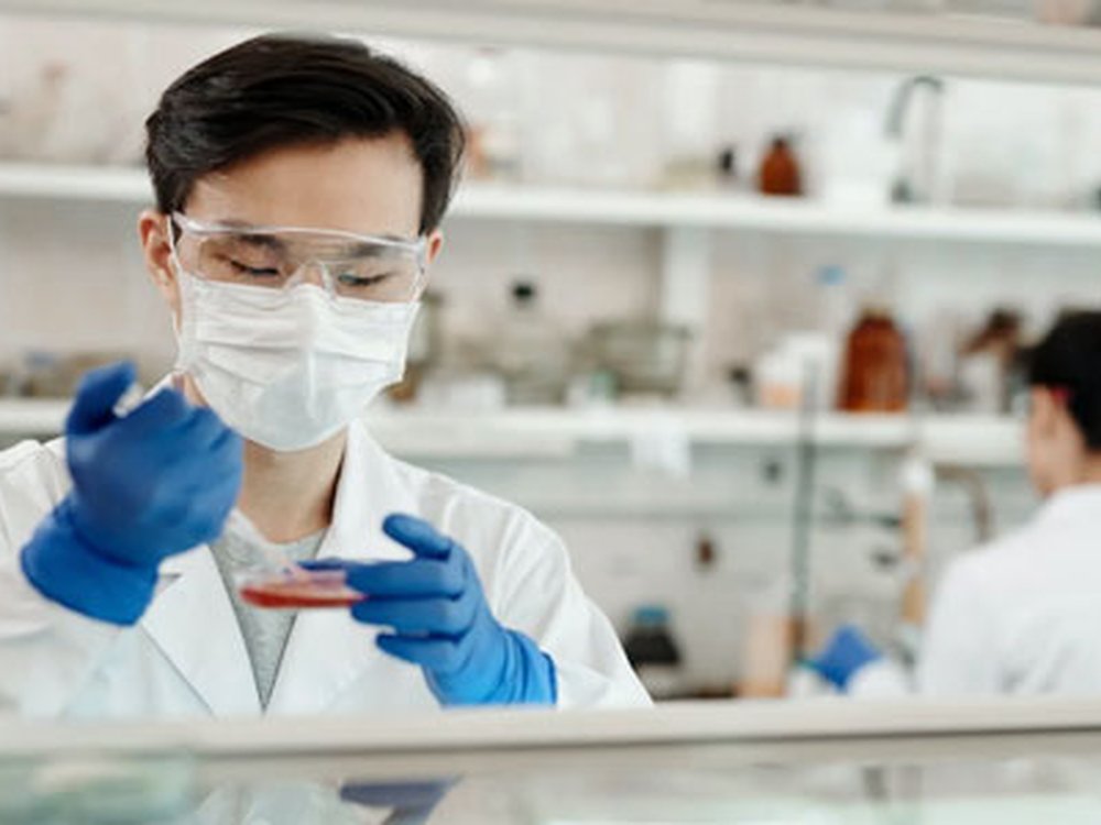 Working for a better future for people with mito - A man in a science lab wearing a white coat, face mask and protective blue gloves using scientific equipment