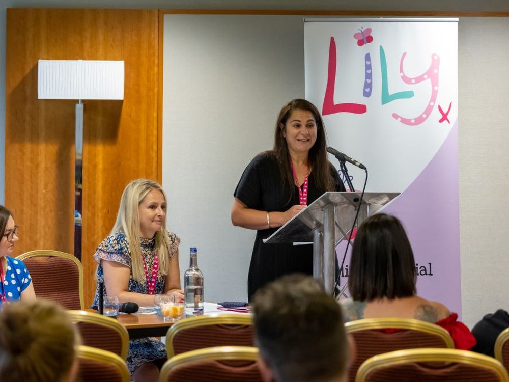 Our commitment to finding treatments and a cure - Lily founder Liz standing at the front of a room talking to people seated in front of her with a Lily Foundation banner behind her