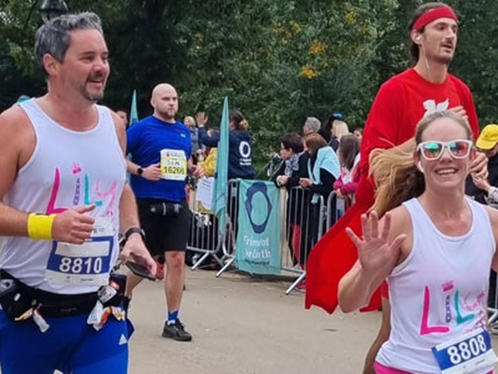 Amazing Lily Events to challenge and inspire you - A man and a lady wearing white Lily vests and running in an official race with other runners around and spectators at the side of the road behind barriers