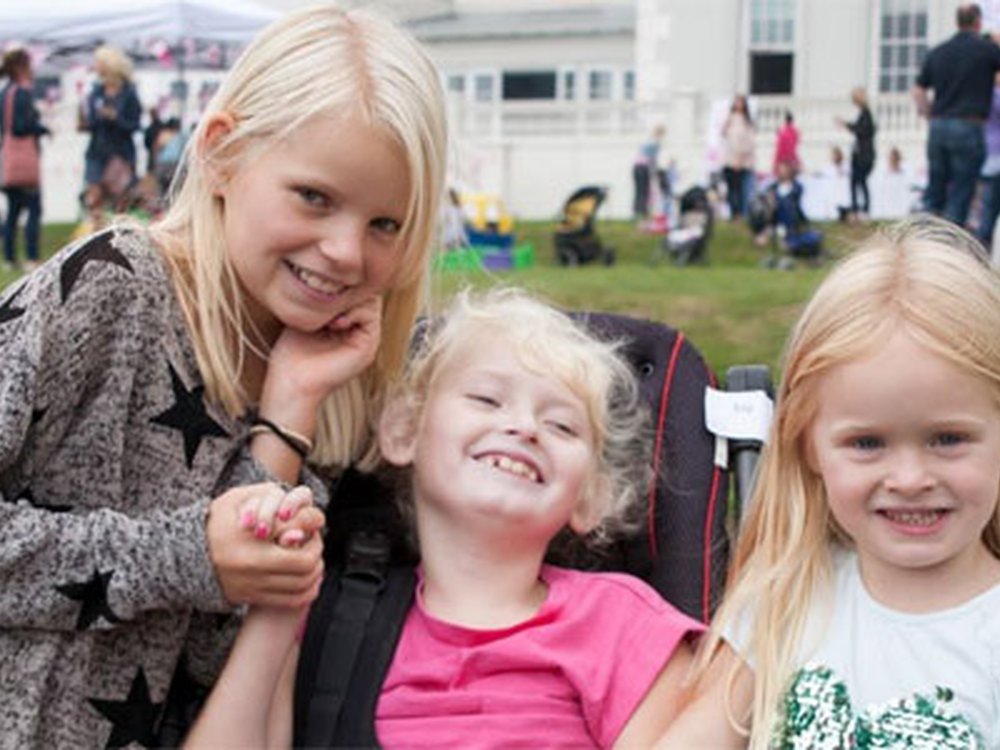 Make your kindness count with Lily Regular Giving - A young mito warrior in her wheelchair with two girls standing either side of her smiling