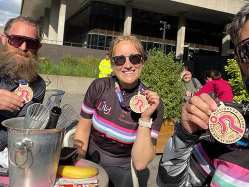 Sign up and get mito-vated! - Three cyclists in Lily tops sitting around a table holding up their RideLondon medals in the sunshine