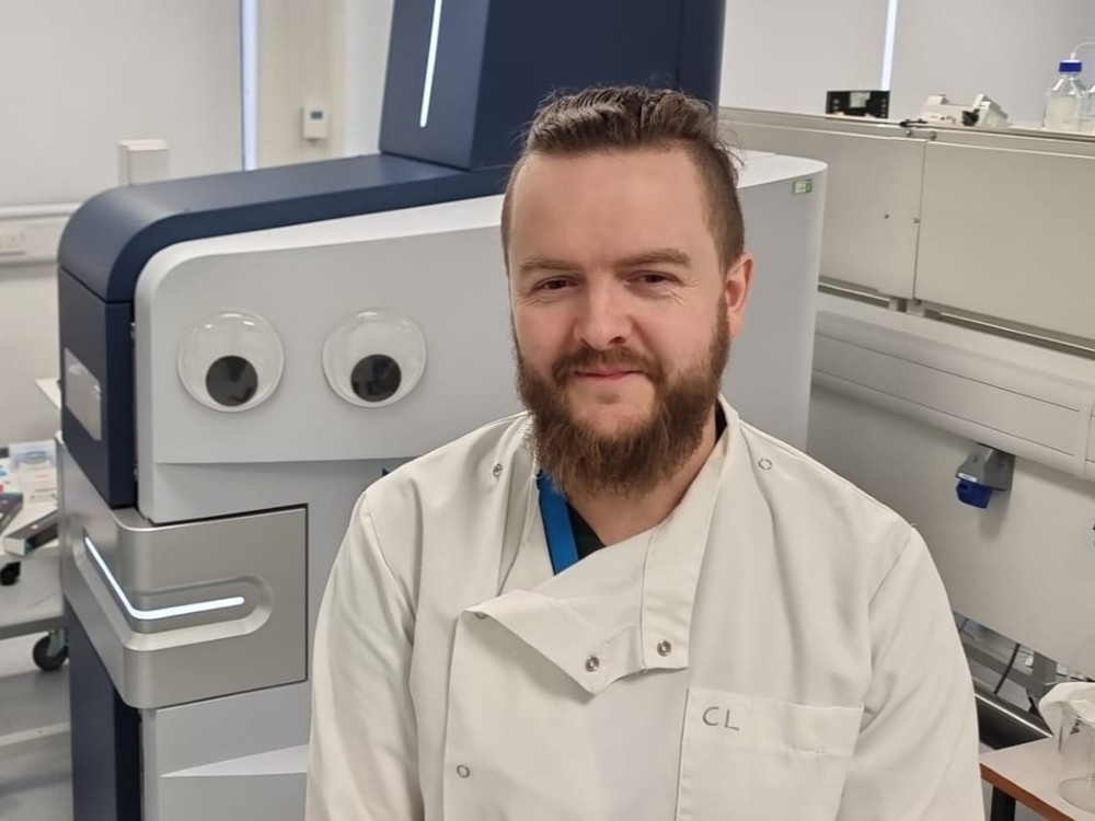 A man standing in a lab in a white coat with a large white machine behind him