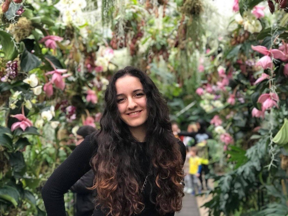 A lady with long dark hair standing in front of lots of greenery smiling