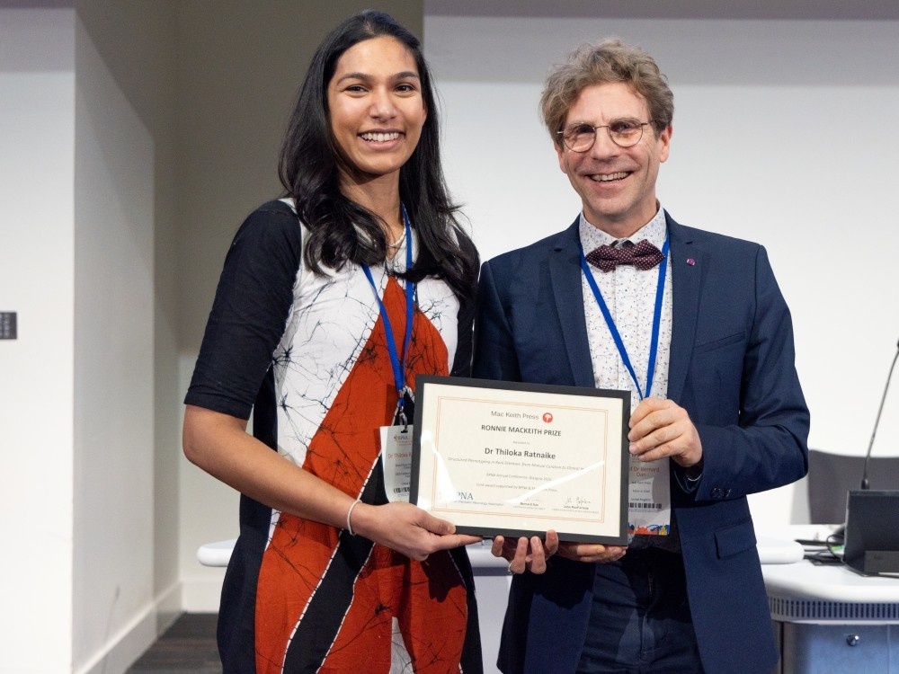 A woman and a man stand together smiling, each holding a framed prize certificate