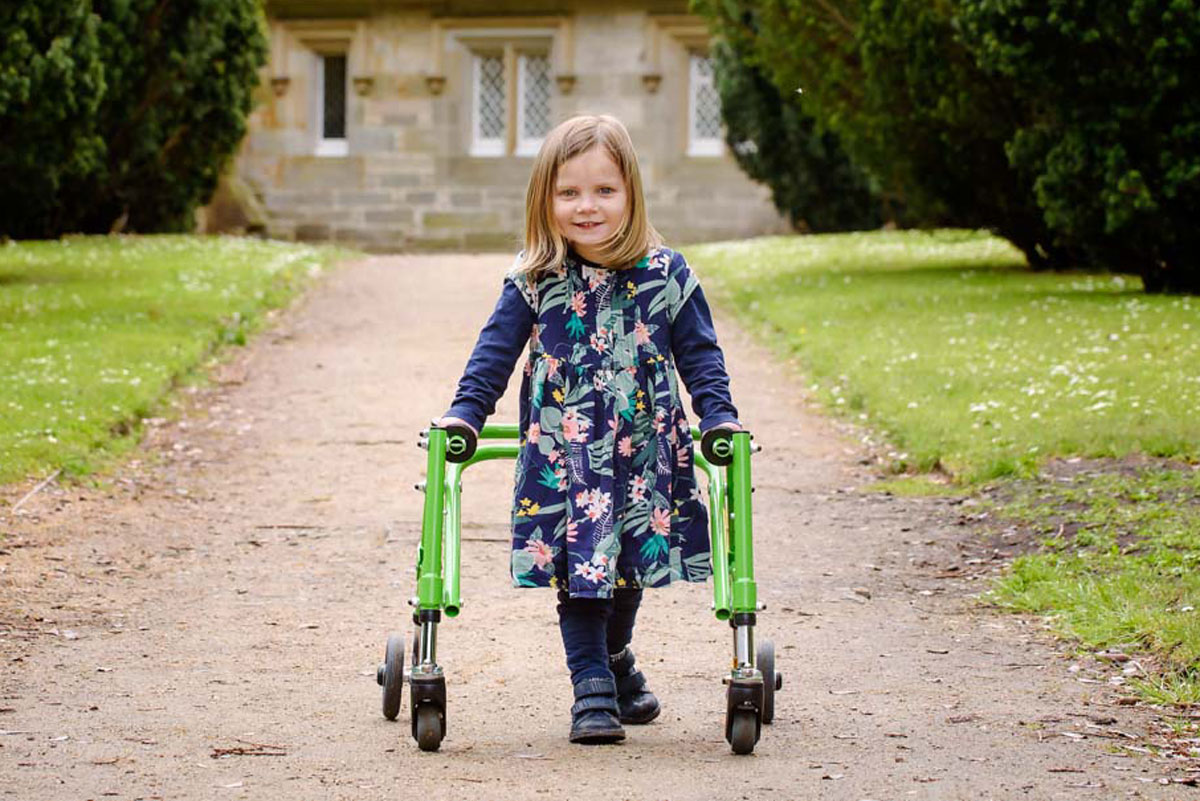 A little girl with mitochondrial disease walks towards the camera in a walking frame