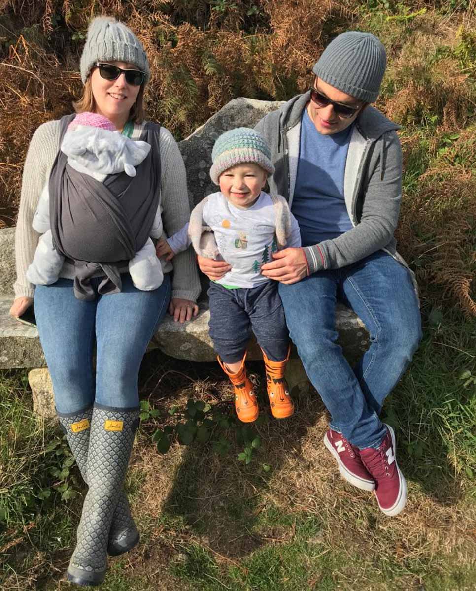 A man and women in woolly hats sit on a bench, between them is a little boy with a woolly hat. The women has a baby attached to her front in a sling