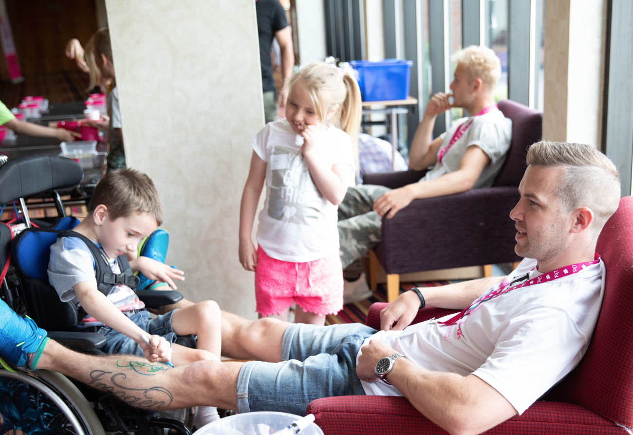 8-year-old James Warwick Dodd, who has mitochondrial disease, with volunteer Neil Harper at a Lily Foundation event A small boy in a wheel chair draws on the leg of a man sat in a red chair with his feet up on the wheels of the wheelchair