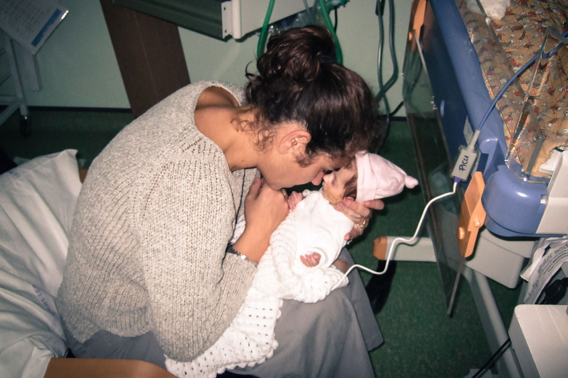 A woman sitting in a chair holding a very small baby in a pink hat. Their noses are touching and eyes are closed. The baby is attached by wires to a medical device