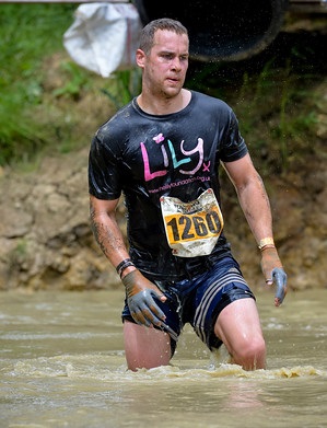 A soaking wet man in a black Lily foundation tshirt with a race number 1260 pinned to his front walks through a pool of muddy water that is up to his knees