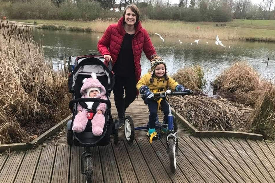 A women stands on a pontoon next to a river. One side of her is a baby in a pram and the other side is a little boy on a trike. Everyone is dressed in warm clothing